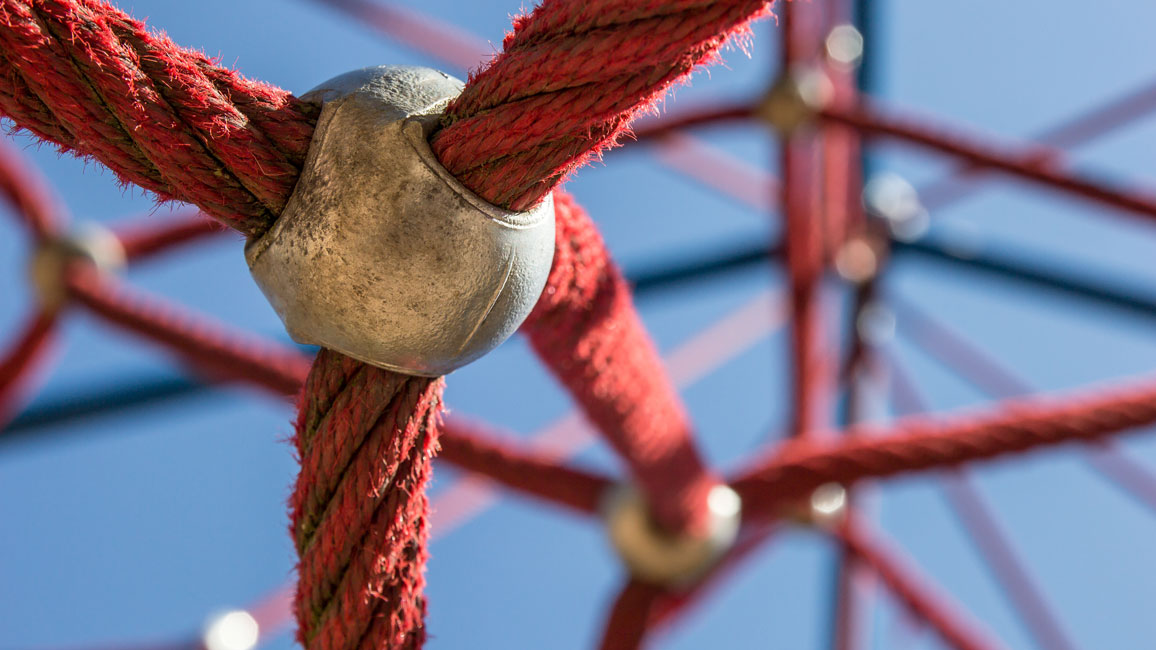 Detailansicht eines Klettergerüsts: Rote Seile sind miteinander durch runde Metallkugeln verbunden, erinnernd an ein Netzwerk.